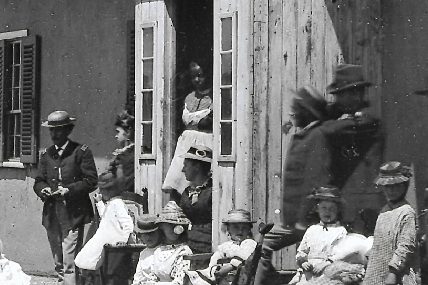 A group of people stand outside Fort Garland adobe building. A young Black woman stands inside of a doorway.
