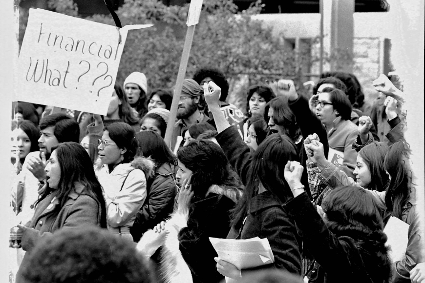 The image depicts a group of students and other supporters at the University of Colorado Boulder protesting the lack of financial aid for Chicano/a students. One student holds a sign that reads "Financial What???" Several other students hold their fists in the air.