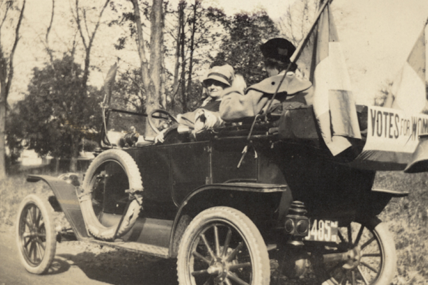 Suffragettes hang flags with "Votes for Women" on the back of an old automobile.