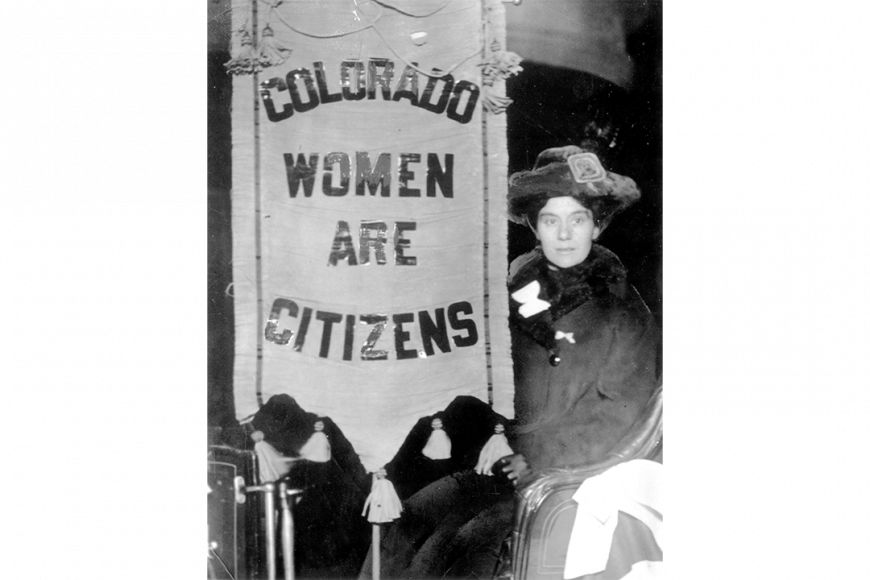 A young woman suffragette sits in a chair near a banner reading "Colorado Women are Citizens." The woman wears a fur coat, hat, and leather gloves. Tassels dangle from the top and bottom of the banner.