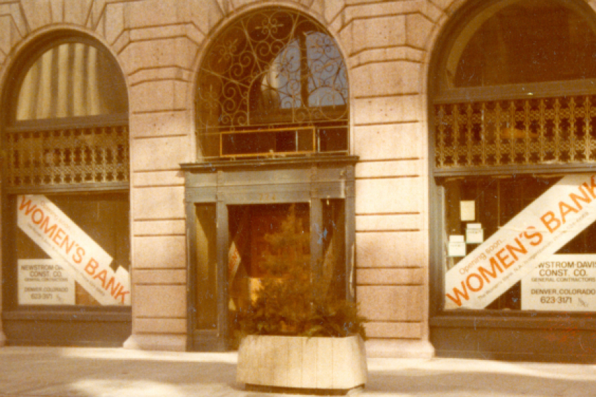 Exterior of arched brownstone building, on the front are two large white signs with red text that reads "Women's Bank"