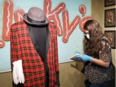 Photo of Kristin Chiesi, completing a condition report on Blinky the Clown's Jacket before taking it into History Colorado's collection. Kristin is seen here, standing in front of a mannequin on which Blinky's iconic red plaid jacket, black bowler hat, and white gloves are positioned. Kristin is wearing blue latex gloves and holds a clipboard. Because of COVID, she is also wearing a face mask.
