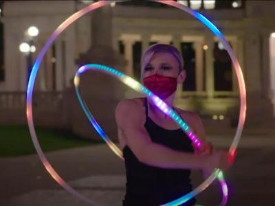Photo of a woman in the middle of a hula hoop performance. She is using two hoops of different sizes, which are illuminated by multicolored LED lights. She is wearing a black sleeveless leotard and she is wearing a mask as a coronavirus precaution.