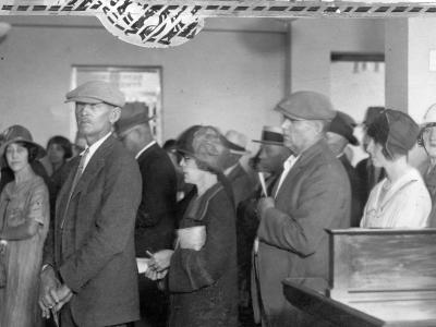 A photo of a queue of customers at Broadway National Bank in Denver. A man is conducting business at the teller window to the left of the image, while several people are waiting quietly in line behind him, many holding a handbag or documents.