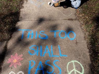 Photo of a city sidewalk, flanked by grass on each side. A child sits on the edge of the grass toward the top of the photo, and they are holding sidewalk chalks. A bucket of chalks is on the ground next to their foot, and they are wearing jeans, a sweatshirt, and a hat. The sidewalk is decorated with hearts and flowers in yellows, blues, and pinks, along with yellow peace symbols. In large blue letters are the words "This too shall pass."