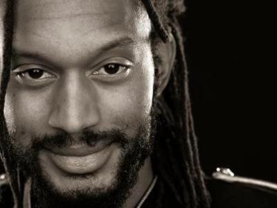 Photo of African American poet Stephen Brackett. Photo is a close up of his face and part of his left shoulder.  He has long hair in dreadlocks, and a shorter beard and moustache, and a warm, friendly expression on his face.