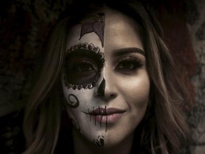 Photo of a young woman with long hair. One half of her face is painted as a white skull, with decorative thin black scrolls on her right cheek and a large black circle around her right eye. The eye circle is lined with gold sequins. She also has four thin black vertical lines across her lips on the right side of her mouth.