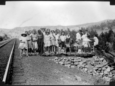 A group of African American teenage campers and their counselors from Camp Nizhoni pose by Denver & Rio Grande Western Railroad tracks and a railway stop near the entrance to Lincoln Hills (Gilpin County), Colorado. They wear dresses, blouses, shorts, and wide-brimmed hats; some hold jackets and coats. Shows luggage on a wooden platform.