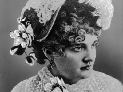 Image portrait of Baby Doe Tabor from the shoulders up. She is wearing a white lacy blouse with a flower attached on the right side of the high collar. She is wearing a very fancy hat on her head, that has white flowers on the right side of it and a large white ostrich feather on top. Her curly hair is seen beneath the hat, and she wears dangling earrings made of many small pearly beads.