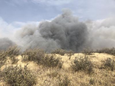 Photo of a massive dark gray wall of smoke rising up just beyond the hilltop in the foreground. The hill is covered with brown grasses and low shrubs. The sky over the billowing smoke is bright blue.