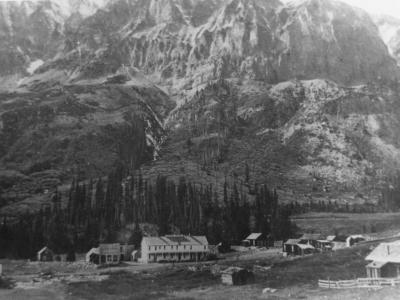 Photo of the town of Gothic, Colorado, dating from the 1880s. Buildings stand at the foot of a rocky mountain which has been dusted with snow. Most of the buildings are small one-story cabins, and one building in the center of the photo is a large white two-story building.