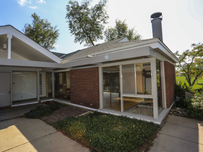 Photo of a midcentury home in Denver. The one-story building is made of red brick with white trim and windows which make up partial sections of exterior walls of the home. The windows are free of visible interior window coverings, and a few minimal pieces of furniture can be seen indoors. The home is landscaped using low-lying green shrubbery, laid brick walkways, and a large concrete pad in front of the home. The front door is large and white, with only a lock, knob, and mail slot decorate the door.