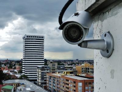 Photo of a security camera attached to the exterior wall of a high-rise building in a city. The security camera is looking directly at the viewer of this image.