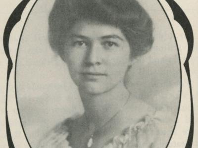Photo of a young woman, posing for a portrait. She wears a light colored blouse with small ruffles around the neckline, and a pendant necklace. She is fair-skinned with dark hair  that is pulled up into a bun at the top of her head. She has a faint smile on her face.
