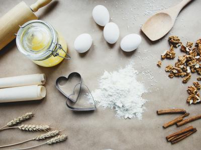 Photo of ingredients used in baking, laid out on a counter. A wooden rolling pin and a roll of parchment, along with a few sheafs of wheat are on the left of the image, while a small mound of flour is next to 2 heart-shaped cookie cutters in the center. Four eggs are nearby, as well as walnuts and stick cinnamon.