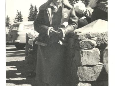 Photo of a woman standing next to a pillar of stacked stones, her left elbow leaning lightly against the top of the stack. A few other people are nearby, facing in the opposite direction. The woman is wearing a below-knee skirt with a blouse and buttoned short jacket, over which she has a leather bomber-style coat. She is wearing a hat on the back of her head, under which her hair is tucked. She is smiling slightly, looking at the person taking the photo.