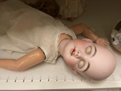 Photo of a doll, laying on a metal shelf lined with a thin sheet of plastic packing. The doll is dressed only in a cotton underdress with no sleeves. The doll has no hair and its eyes are closed, but because of the difference in color of eyelid and face, it appears as though the doll has blank eyes. Its orange-lipped mouth is open, and its arms are askew.