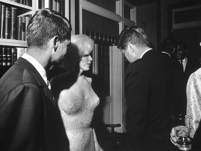 Photo of Robert Kennedy on the left of the image, standing next to Marilyn Monroe who is wearing the iconic nude-looking dress that is covered in rhinestones. President Kennedy stands next to her with his back to the camera and they are in a room with shelves filled with books. There is a partial image of a man captured on the right edge of the image. They are talking and smiling.