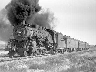 Black and white photograph of steam engine billowing black smoke and hauling a train of cars.