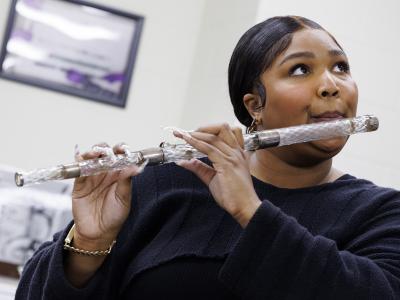 Photo of pop performer Lizzo, holding a flute to her mouth to play. The flute is crystal with silver bands. Lizzo is a Black woman, whose black hair is parted in the center and smoothed, pulled to the back of her head. She is wearing gold earrings and a bracelet, and a navy blue long sleeve sweater. She is standing in a bright white room that has a framed image hanging on the wall behind her. She is looking up toward the ceiling as she plays, her lips pursed as she blows into the instrument.
