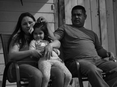 A family sits on a porch. A man sits on a chair to the right, while a woman sits on a chair to the left with a young girl in her lap.