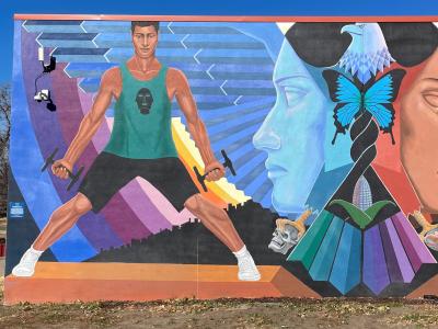 A colorful mural depicting an eagle whose wings form the faces of Chicana women. On one side is a modern chicano athlete, on the other side is an Indigenous person participating in a ritual.