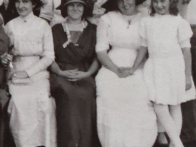 Three women in turn-of-the-century dress sit together in an outdoor setting. The second from the left is wearing a dark dress, while the other three wear light colored or white dresses. The three older women are wearing broad-brimmed hats.