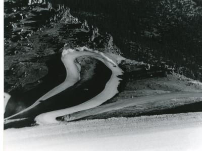 Photo of a portion of the twists and turns of the Pikes Peak International Hill Climb. The view is of a race vehicle having just gone around a hairpin turn, kicking up a trail of dust from the dirt track. Beyond the track lies the rocky hill top of the mountain, the steep terrain evident as the car climbs higher along the course.