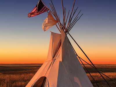 A tipi at sunset. Over the tipi fly two flags: the flag of the United States, and the white flag of peace.