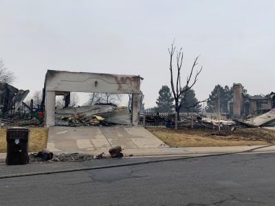 A burned house sits on a street. Only one front wall is left partially standing, and through the gaping garage door a pile of rubble is visible.