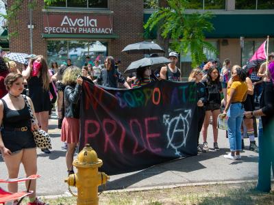 Photo of activists standing in front of a brick building. There is a group of a few dozen people in this image, standing in small groups talking with each other, looking around. A few people in the front carry a sign made of black fabric. The sign says "NO CORPORATE PRIDE" painted in colorful letters, and a large transgender symbol (a pictogram consisting of a circle with an arrow sprouting from the top-right side, a cross at the bottom, and a stroked arrow at the top) painted in white in the lower corner.