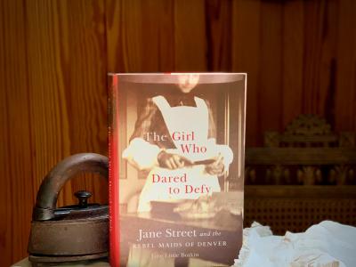 Photo of a book sitting on an ironing board, next to an antique iron and a white garment. Titled "The Girl Who Dared to Defy: Jane Street and the Rebel Maids of Denver," the cover is a black and white image of a female who wears a dark colored dress over which a long white bibbed apron is worn. She is standing in front of a table with a shallow small bowl and what appears to be a rectangular cooking pan. Although only part of her face is seen, she looks down at an open book she holds in her hands.