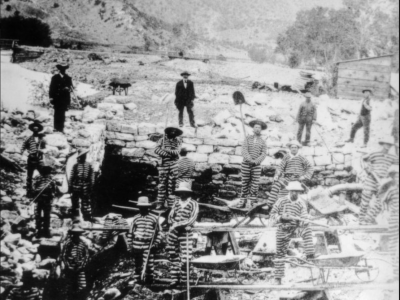 Black and white photo of men in striped prison uniforms digging a large hole. Two guards in dark uniforms observe. A river and mountains are in the background.