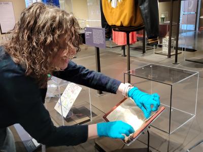 Photo of a member of museum staff, carefully caring for the pages of a document that is on display in the Zoom In exhibition. The staff member wears blue nitril gloves to handle the pages of the document. 