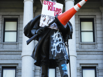Image of a vandalized statue outside of a government building. Signs and graffiti say "Stop killing us" and "ACAB"