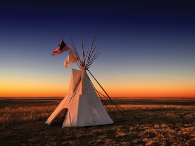 A tipi at sunset. Over the tipi fly two flags: the flag of the United States, and the white flag of peace.