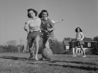 Black and white image of three young women kicking a ball in a grass field. There's houses and two adults behind them.