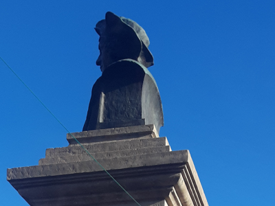 Profile view of a dark stone statue depicting the bust of Christopher Columbus. The American flag waives behind it.