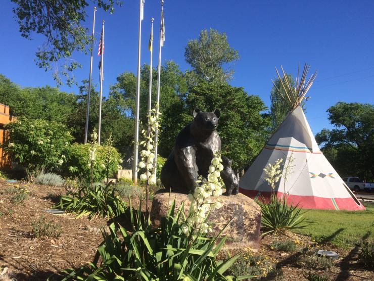 The grounds of the Ute Indian Museum. A tipi is visible in the background, along with several trees and flagpoles. In the foreground are smaller plants, as well as a metallic statue of a mother bear and cub.