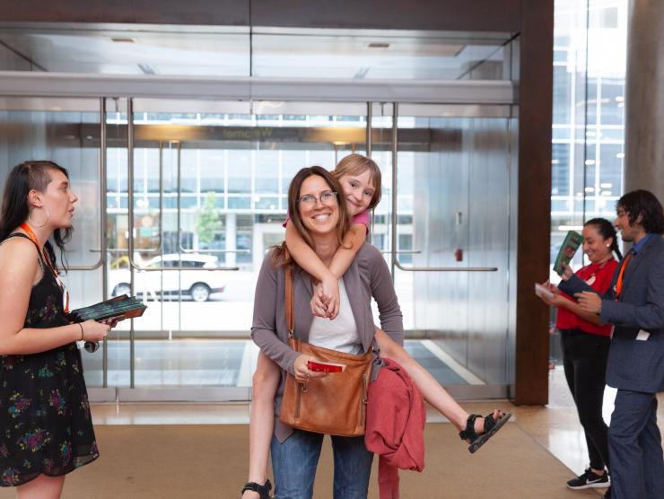 A History Colorado member and her daughter enter the History Colorado Center, smiling. The child is riding on her mother's back.