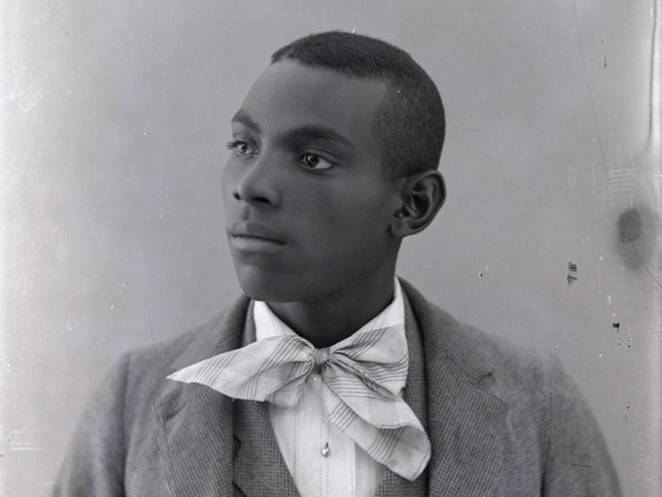 The image depicts a studio portrait of a young man. Only his bust and head are visible in the portrait. He is posed with his head looking to the left. The young man wears a lightly colored checkered three-piece suit with a white pin-stripe button down shirt. He also has on a plaid neckerchief tied into a bow.