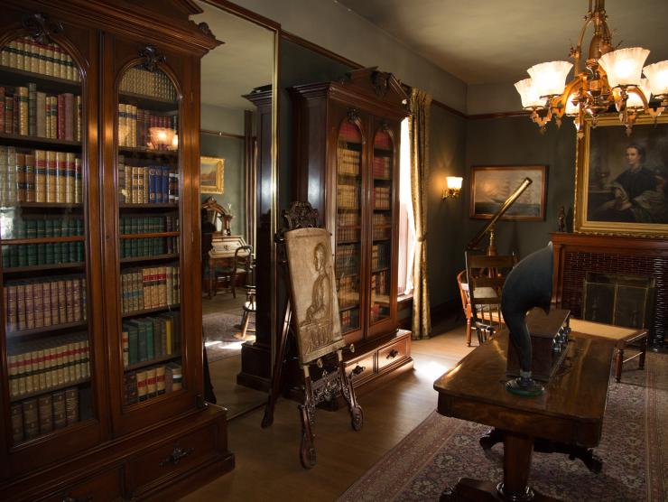 The library at the center for Colorado women's history. The dark wooden bookshelves are lined with old books. There is a record player on the table and an old portrait behind it on a dark green wall.