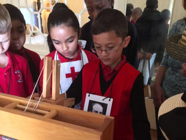 A group of children wearing red shirts huddle around an antique loom.