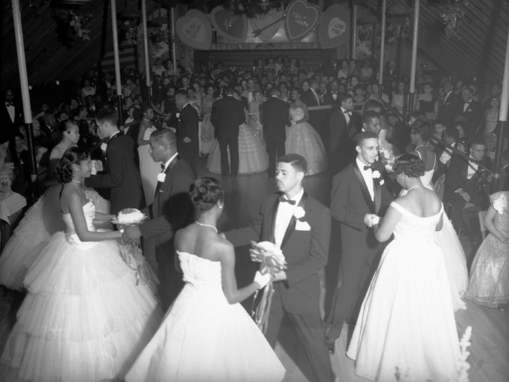 View of an Owl Club function. A group of African American (Black) teenagers stand in a circle on a ballroom floor in Denver, Colorado. The young women wear formal dresses and the young men wear tuxedos.