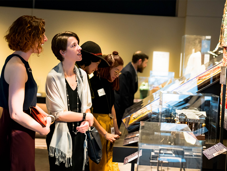 A group of people standing in front of display cases at our exhibition, Zoom In. 2 women with short hair in black dresses speak.