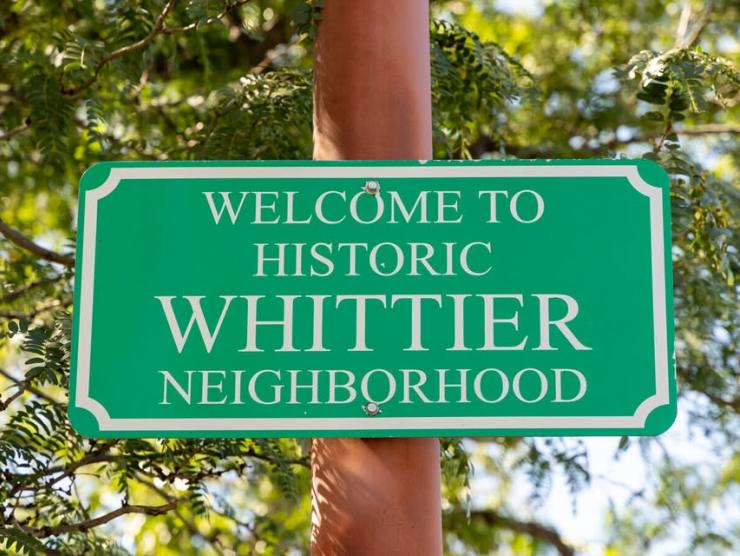 "Welcome to historic Whittier neighborhood" on a green street sign attached to a wooden lamp post. Behind it are branches of evergreen trees.