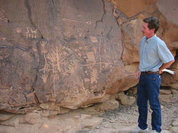 A tour member stands and admires a rock wall covered in large petroglyphs.