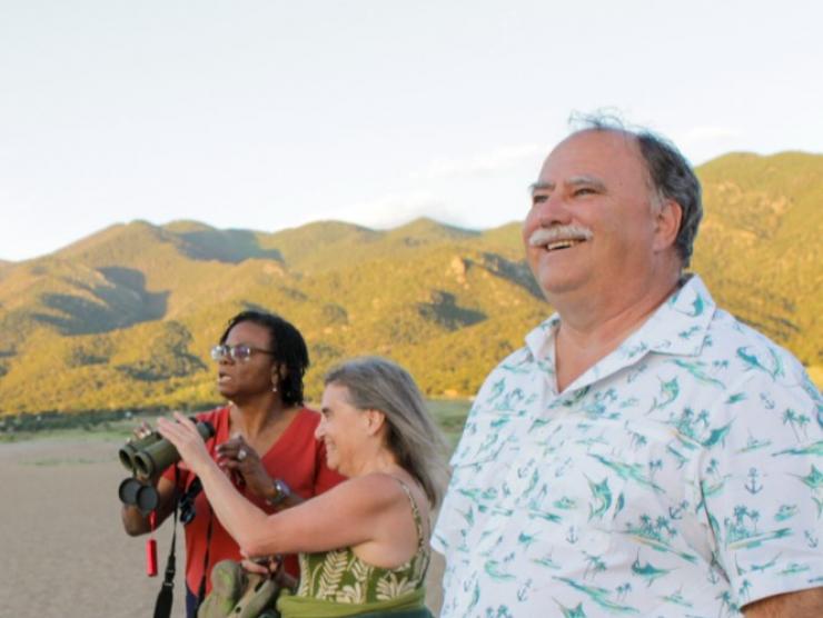 An older gentleman smiles at the Sand Dunes vista with mountains behind him. In front of him, two women share binoculars and enjoy the view.