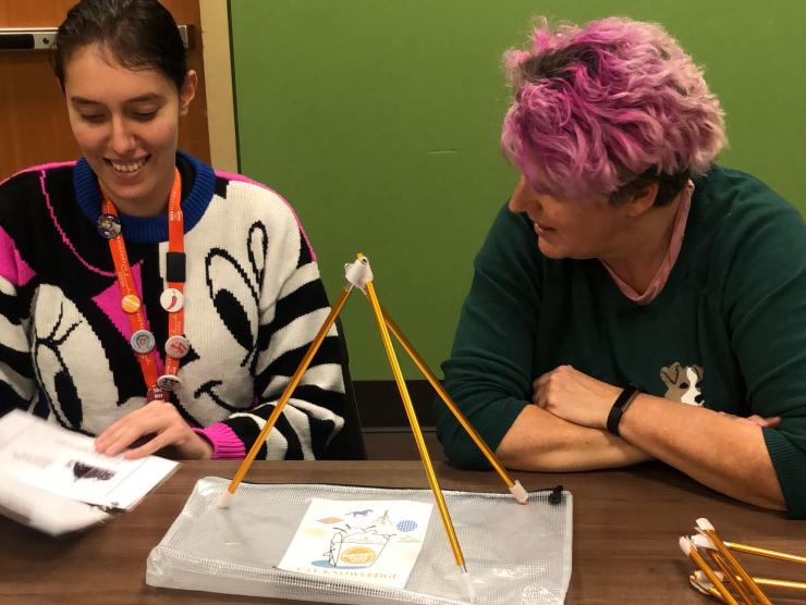 2 teachers hold papers and balance sticks into a pyramid while working on curriculum planning and professional development together.