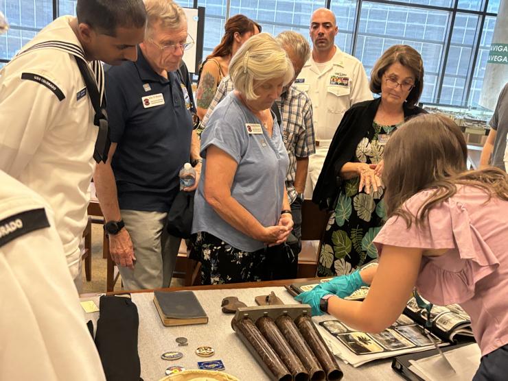 History Colorado staff demonstrates with a number of artifacts at a table while a crowd of guests including members of the USS Colorado, look on.